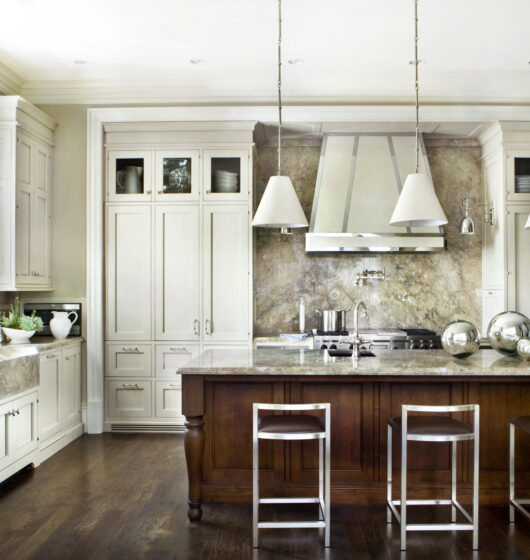 A modern kitchen with a large marble-topped island, three metal bar stools, and pendant lights. Stainless steel appliances, white cabinets, and marble backsplash complete the elegant look, with natural light streaming through a window.