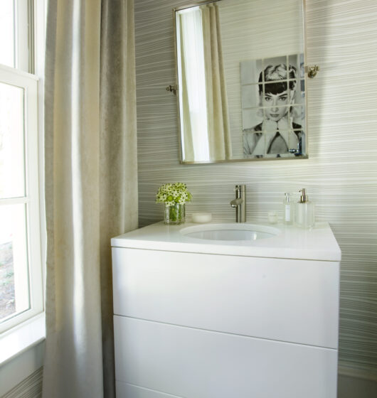 A modern bathroom features a white vanity with a sink, a square mirror above, and toiletries. Vertical striped wallpaper and a window with a long curtain add elegance, while a monochrome artwork hangs on the wall. A small plant sits on the counter.