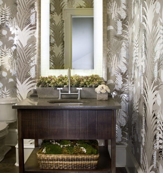 Bathroom with a minimalist design featuring a backlit mirror above a sleek sink on a wooden vanity. The walls are covered in patterned wallpaper with a nature motif. A basket with greenery sits on a shelf below the sink.