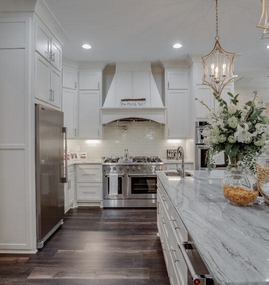 Luxurious kitchen with white cabinets and a marble island. Stainless steel appliances, including a double oven, are featured. Elegant hanging lights illuminate the space. A vase with white flowers and a bowl of cereal are on the island.