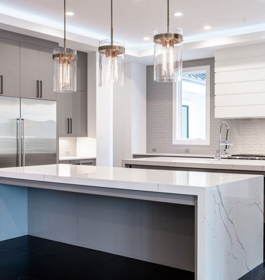 A modern kitchen with a large marble island, stainless steel appliances, dark wood flooring, and sleek gray cabinets. Pendant lights hang above the island, and a window provides natural light in the background.