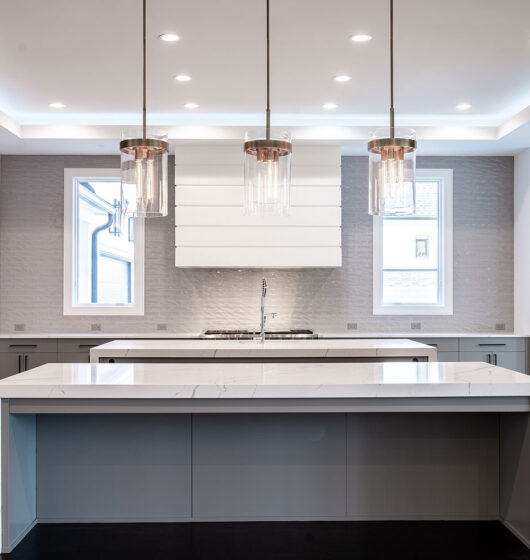 Modern kitchen with a large island and marble countertop. Three pendant lights hang above. White cabinets and stainless steel appliances are present, with two large windows providing natural light. Wood flooring adds warmth to the space.