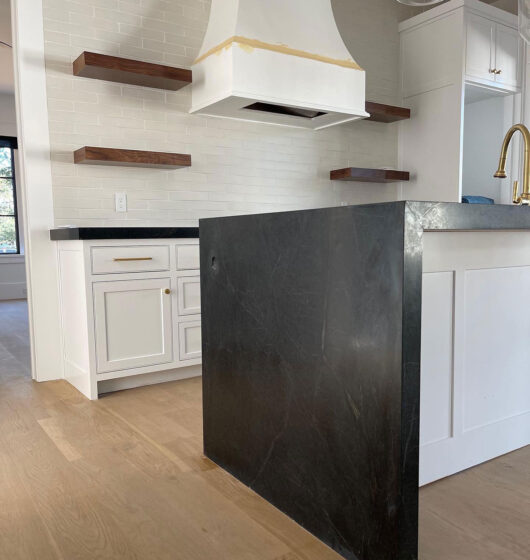 Modern kitchen with white cabinets, a black countertop with a waterfall edge, and wooden floating shelves against a white tile backsplash. A brass faucet is visible. Light wood flooring adds warmth to the space.