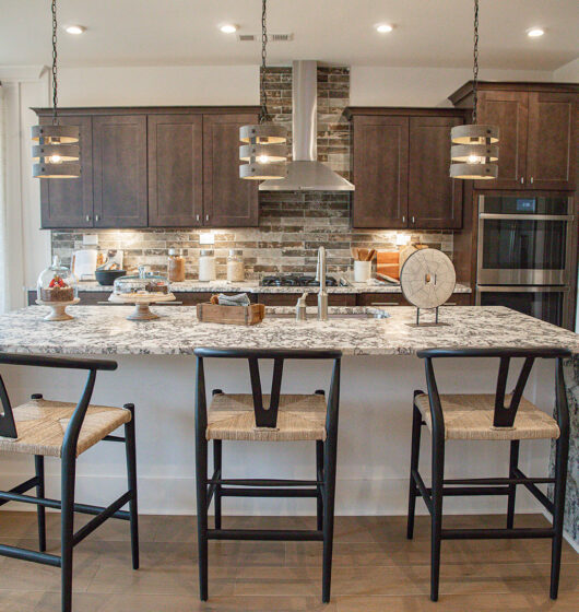 Modern kitchen featuring a large granite island with three wooden barstools, two pendant lights above, dark wooden cabinets, a stainless steel oven and refrigerator, and a decorative brick backsplash. The room is bright with natural light.