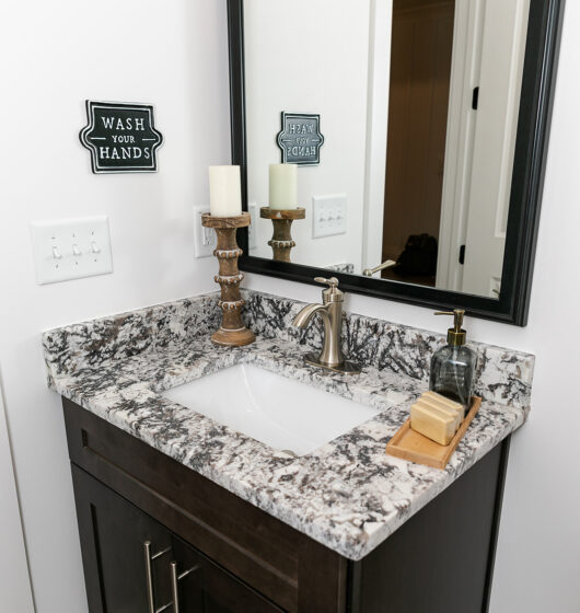 A bathroom vanity with a dark wood cabinet, marble countertop, and a rectangular mirror. The sink has a bronze faucet. A soap dispenser and candleholder sit on the counter. Signs on the wall read Wash Your Hands and Dream Soap.