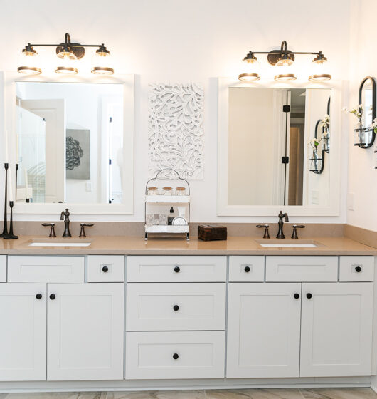 Bright bathroom with a double vanity featuring two sinks, bronze fixtures, white cabinetry, and large mirrors. Two sets of wall lights illuminate above the sinks. Decorative white wall art is centered, with towel rings on the right wall.