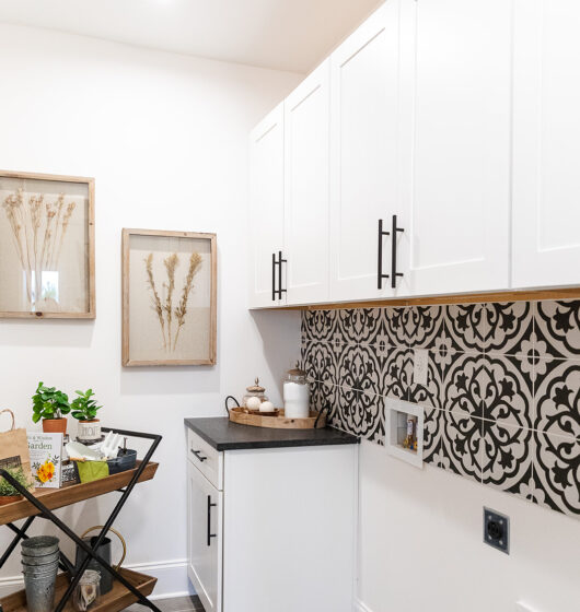 A modern kitchen corner features white cabinets with black handles, a black and white patterned backsplash, and gray tile flooring. A cart holds plants and a wooden tray with a mug. Two framed botanical artworks hang on the wall.