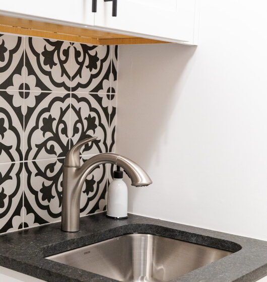 Modern kitchen sink with a stainless steel faucet set in a dark countertop. White cabinets are above, and a black and white patterned backsplash adds contrast. A soap dispenser is placed near the faucet.