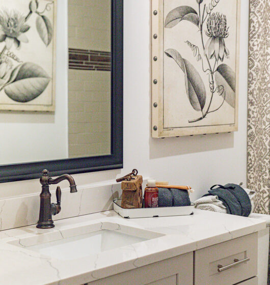 A bathroom with a white marble countertop and a black faucet. A mirror reflects a botanical wall art, which is also visible on an adjacent wall. A tray holds grooming items and two rolled towels. The decor includes a patterned curtain.