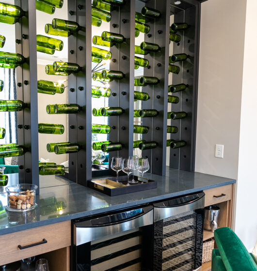 Home wine bar with a modern design, featuring green wine bottles on a vertical display. The countertop holds several wine glasses, and there are dual wine coolers below, with wooden cabinets and baskets. A glass container with corks is on the counter.