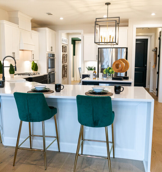 A modern kitchen featuring a large white island with three green upholstered stools, a gold light fixture above, stainless steel appliances, and white cabinetry. The countertops have black cups and plates, with decorative greenery in the background.