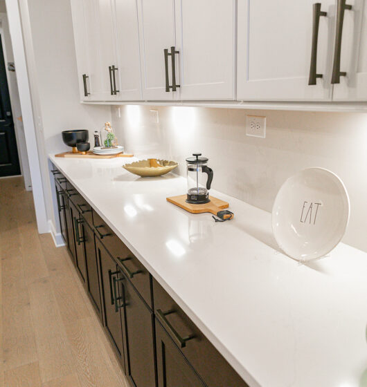 A modern kitchen with white upper cabinets and dark lower cabinets. The countertop is white, featuring a French press, a decorative plate with EAT, and various kitchen items. Hardwood floors and bright lighting illuminate the space.