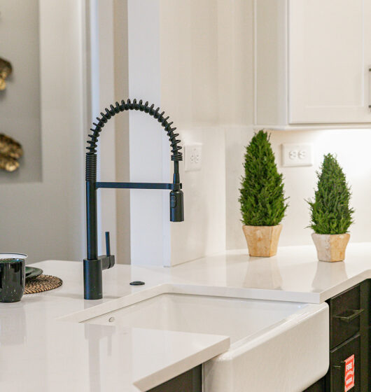 A modern kitchen features a white countertop with a farmhouse sink and black pull-down faucet. Two potted plants are displayed on the counter, and the cabinets and walls are painted white. Bright lighting adds a clean, minimalist feel.