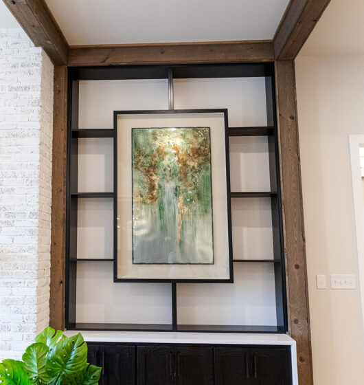 A framed abstract painting with green and gold tones is centrally displayed on a black metal shelf with wood accents. Below, there are dark cabinets. A potted plant with large green leaves sits on a wooden floor to the left.