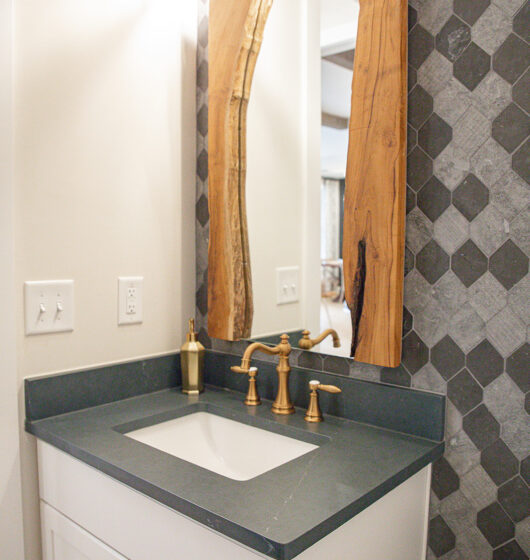 A bathroom vanity with a white cabinet and a gray countertop featuring a sink. It has a unique wooden-framed mirror above and a brass faucet. The wall has gray hexagonal tile accents and there are light switches on the left wall.