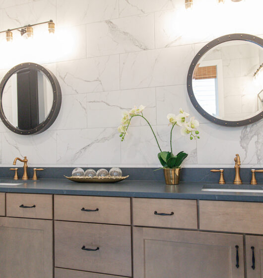 Modern bathroom with marble-tiled walls and a double vanity. Two round mirrors hang above black countertops with gold faucets. A brass planter with white orchids is centered between them, along with a decorative tray. Warm wall lights illuminate the space.