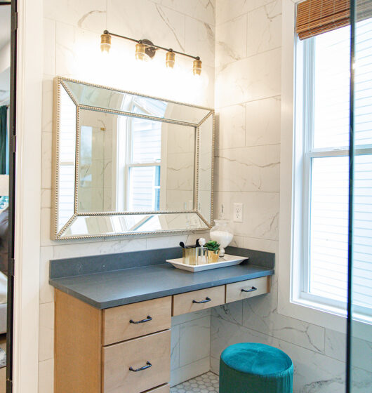 A bathroom with a vanity featuring a large mirror, small drawers, and a black countertop. The wall is tiled in white with a marbled pattern. A round green stool sits on a white hexagonal-tiled floor. A window brings in natural light.