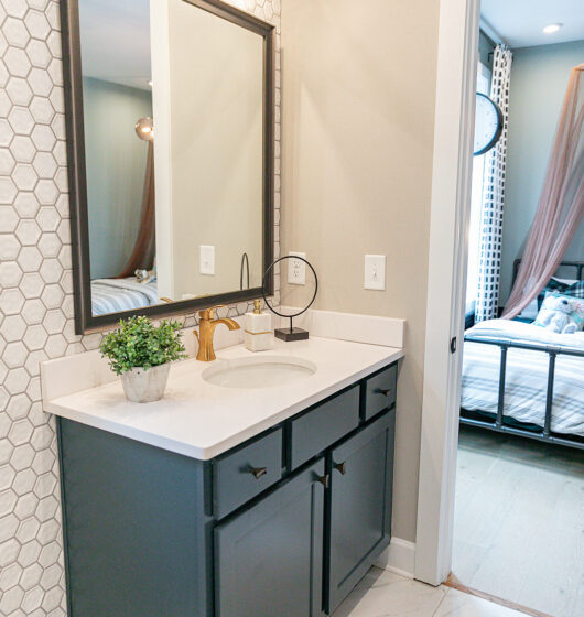 A modern bathroom features a black vanity with a white countertop and a rectangular mirror. A hexagonal tile backsplash adds texture. Gold fixtures, a decorative plant, and a round table lamp accent the space. A glimpse of a bedroom is visible in the background.