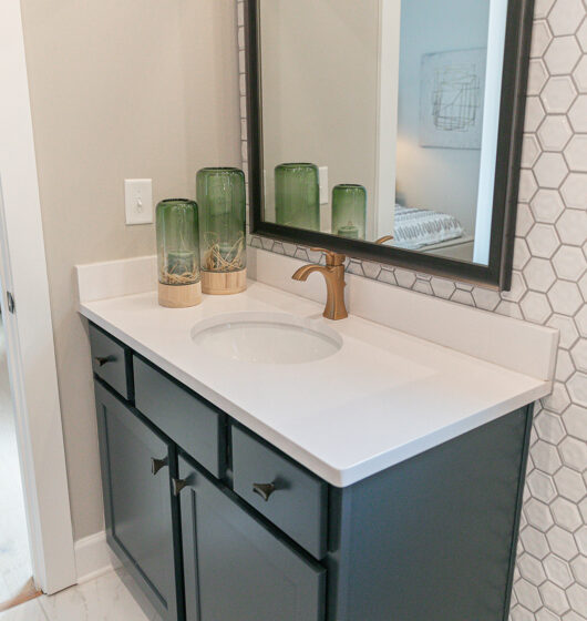 Bathroom vanity with a white countertop and dark cabinets. A modern faucet stands over the sink, and three green glass vases with decorative bases sit to the left. The hexagonal patterned wall tiles add texture, while a large mirror hangs above.