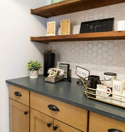 A cozy kitchen nook featuring wooden shelves with decorative items, including signs and potted plants. A vintage rotary phone rests on the dark countertop alongside a tray with cups labeled His and Hers, and a decorative hourglass.