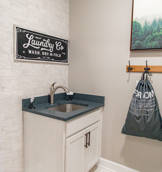 A laundry room with a white cabinet and black countertop. A stainless steel sink is installed on the countertop. Above, a vintage-style sign reads Laundry Co. Wash, Dry & Fold. A bag hangs on a coat rack to the right.