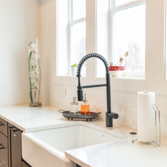 Modern kitchen with a white farmhouse sink, black spring-neck faucet, and soap dispensers. Bright natural light filters through two windows adorned with small plants and an orchid vase. Brown cabinets and a dishwasher complete the clean, minimalist look.