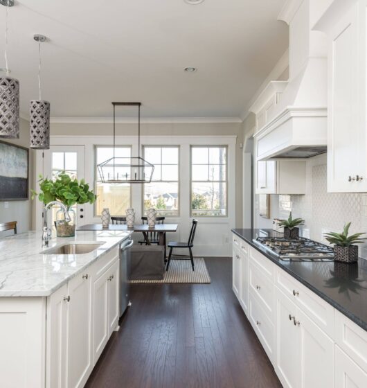 A modern kitchen with white cabinets, a marble island, and dark countertops. Three patterned pendant lights hang overhead. A dining area with a table and chairs is visible, and large windows let in natural light. Decorations include plants and artwork.