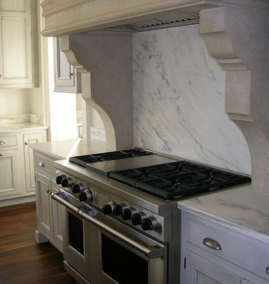 A modern kitchen with a large stainless steel oven and stovetop. The backsplash and countertops are made of white marble, and the cabinetry is a light cream color. The floor is dark wood, adding contrast to the light tones.