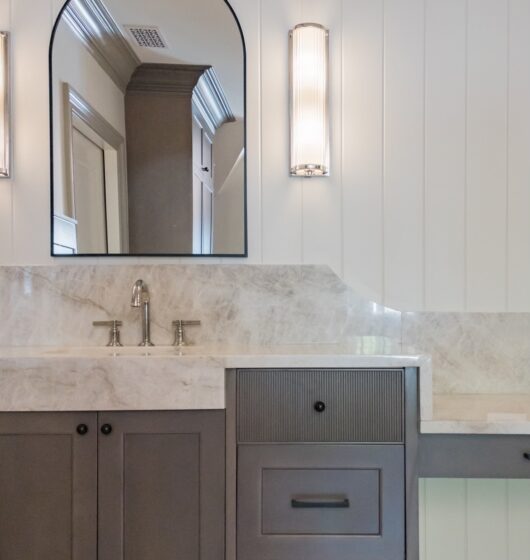 A bathroom features a marble countertop with a modern sink and stainless steel faucet. Above is an arched mirror with vertical light fixtures on each side. The cabinetry is dark gray with a ribbed texture on one drawer front. Light walls and trim.