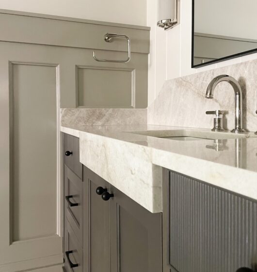 The image depicts a modern bathroom vanity with a textured beige countertop, a stainless steel faucet, and dark cabinet drawers. The wall features wainscoting with a towel ring above, and the countertop curves slightly near the sink.