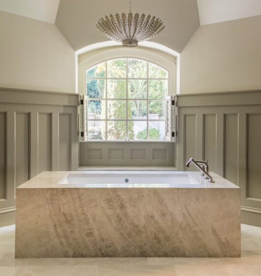 A luxurious bathroom with a large marble bathtub centered under an elegant chandelier. The walls are paneled, and a large, arched window provides natural light, showcasing greenery outside. The floor is tiled in a light, neutral color.