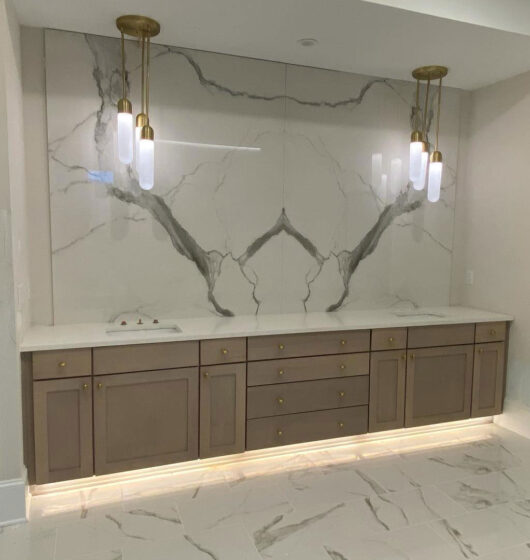 A modern bathroom with a large marble backsplash and beige cabinets. Gold lighting fixtures hang above, and the floor and wall have marble patterns. The area is well-lit, and theres an under-cabinet lighting accent.