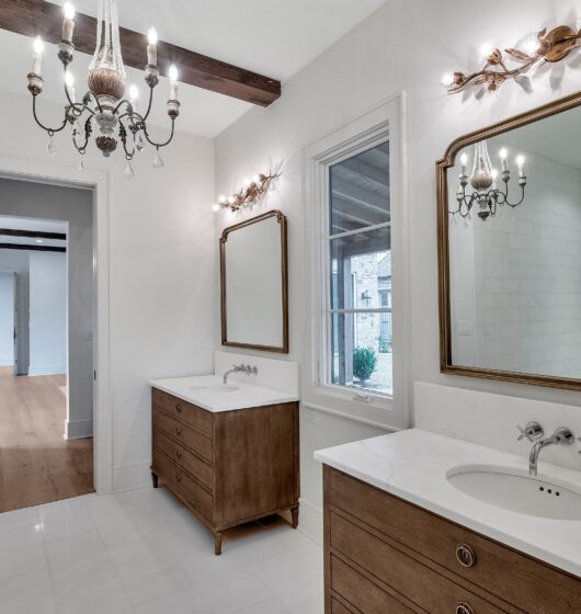 Elegant bathroom with two wood vanities, each with a white countertop and large mirror. A chandelier and ornate light fixtures provide illumination. White walls, a window, and a doorway leading to another room with wood flooring complete the scene.