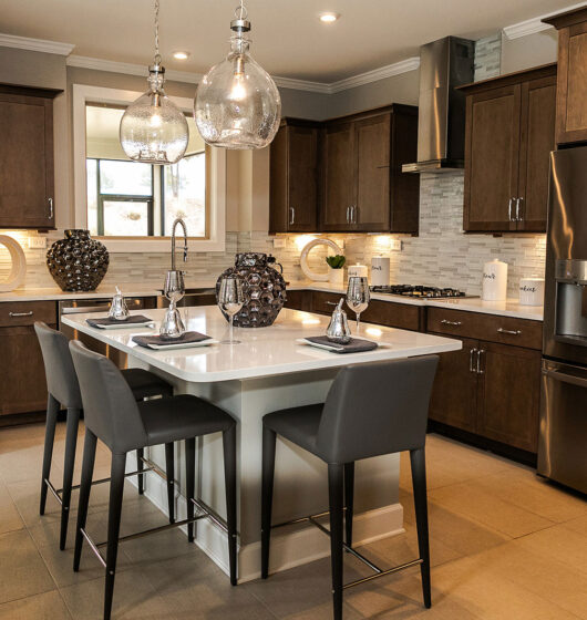 Modern kitchen with dark wood cabinets, stainless steel appliances, and a large island. Three gray chairs surround the island, which is topped with decorative jars and pendant lights. A window and a glass door bring in natural light.