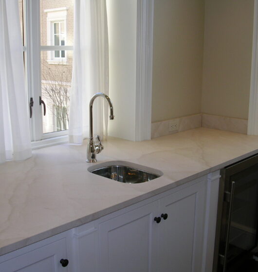 A kitchen corner featuring a marble countertop with a small sink and stainless steel faucet. White cabinets with dark knobs are below. Two large windows with sheer curtains allow natural light. A small wine fridge is visible in the bottom right.