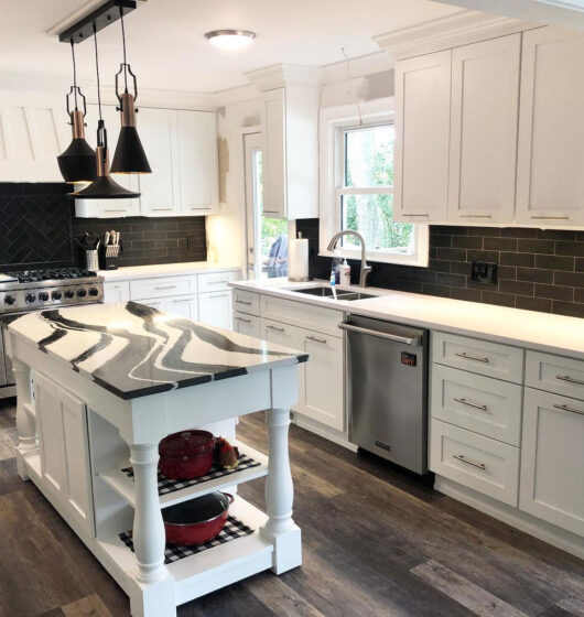 Modern kitchen with white cabinets, a black herringbone backsplash, and stainless steel appliances. A large island with a marble countertop and hanging pendant lights sits centrally. Wooden flooring adds warmth to the contemporary design.