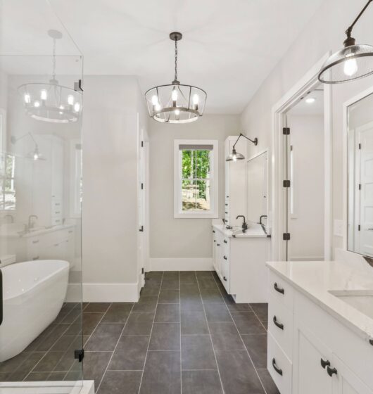 Spacious modern bathroom with a glass shower, freestanding white bathtub, double sink vanity with marble countertop, and large mirrors. Overhead and wall-mounted lights add brightness. Gray floor tiles and white walls enhance the airy feel.