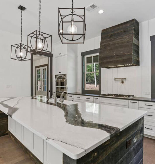 Modern kitchen with a large marble island featuring a unique black and white pattern. Three geometric pendant lights hang above. White cabinets with dark handles, a built-in stove with a wooden hood, and two windows offer a view of greenery outside.