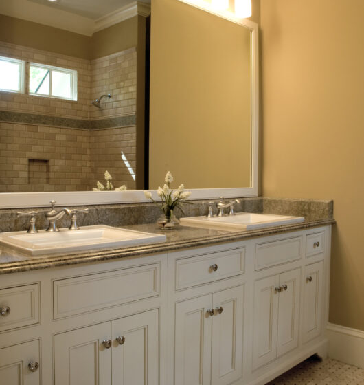 A bathroom with a large mirror above a granite countertop, featuring two sinks. The cabinets are white and have silver handles. Three lights are mounted above the mirror. A vase with flowers sits between the sinks, and a tiled shower is visible.