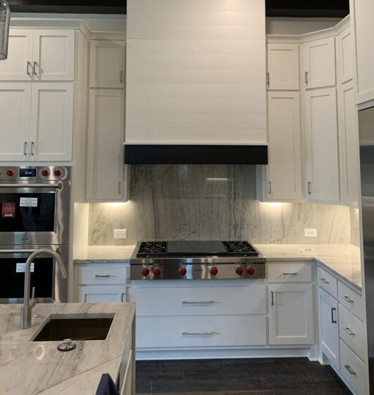 Modern kitchen featuring white cabinetry, a sleek marble backsplash, and a large stainless steel range with red knobs. A double oven is integrated into the left side, and there is a marble countertop with a built-in sink in the foreground.