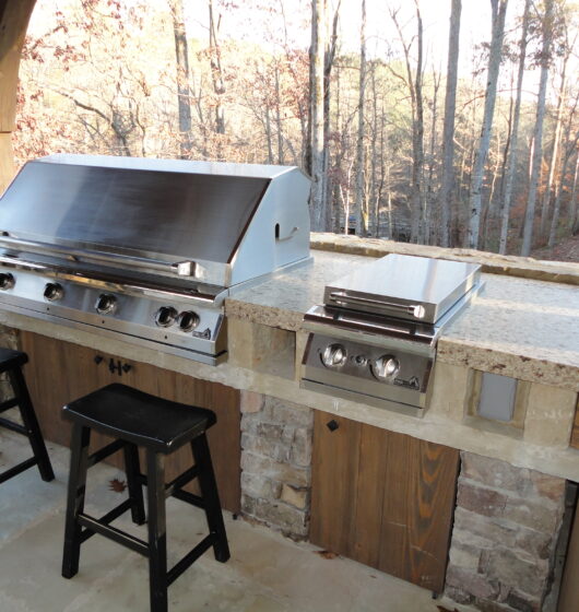 Outdoor kitchen with a stainless steel grill and side burner on a stone counter. Two wooden stools sit in front, and tall trees can be seen in the background, providing a natural setting.