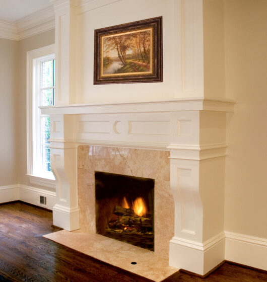 A cozy living room features a large, white mantel fireplace with a lit fire inside. Above the mantel is a framed landscape painting. The room has wooden floors, white walls, and a tall window to the left.