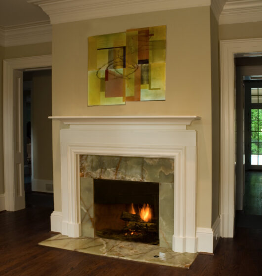 A modern fireplace with a white mantel is set in a light-colored wall. Above it, an abstract painting with yellow and brown tones hangs. The hearth is made of a marble-like material, and theres a small fire burning inside.