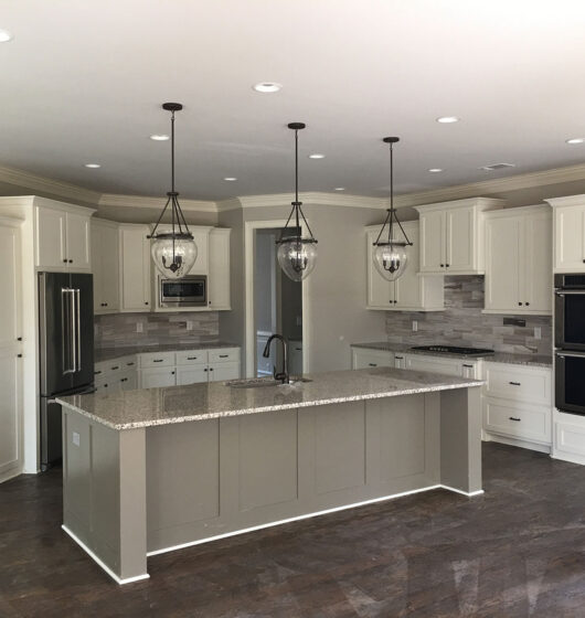 A modern kitchen with a large island featuring a granite countertop, stainless steel sink, and three pendant lights. Surrounding are white cabinets, a double oven, microwave, stainless steel refrigerator, and stone-tile backsplash. Dark wood flooring.