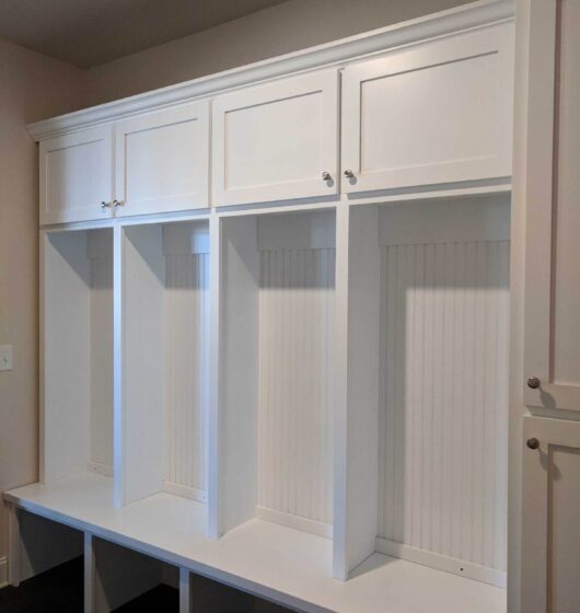 White built-in storage unit with four open cubbies, each with upper and lower compartments. The upper section has a closed cabinet with doors, and the lower section includes a bench. The unit features beadboard backing.