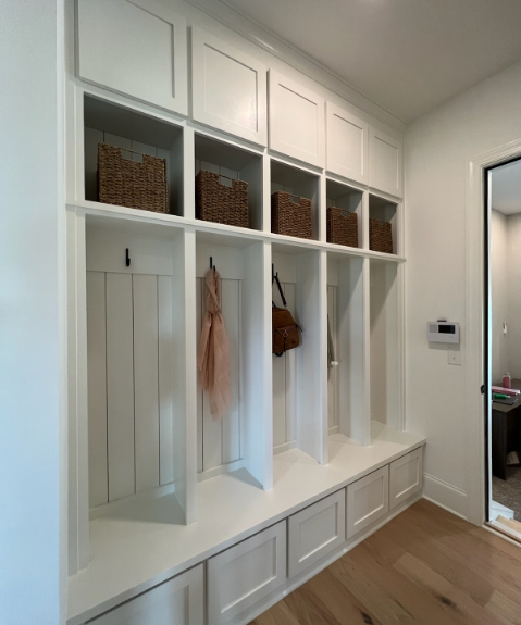 A mudroom with built-in white cabinets and cubbies. Baskets are stored above, with hooks below holding a scarf and handbag. Bench seating offers storage underneath. Light wood flooring enhances the space.