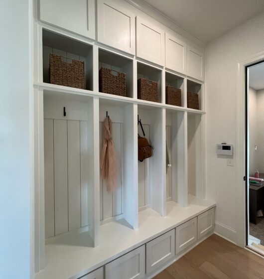 White mudroom with wooden shelves, cubbies, and built-in hooks. Four wicker baskets sit on upper shelves, while hooks below hold a pink scarf and a brown bag. The bench is integrated into the cabinet design, beside a door leading to another room.