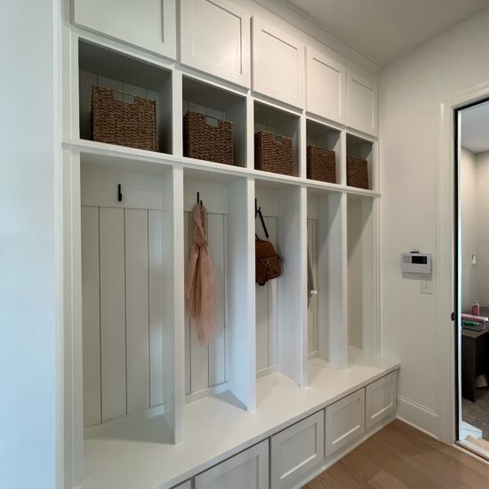 White mudroom with wooden shelves, cubbies, and built-in hooks. Four wicker baskets sit on upper shelves, while hooks below hold a pink scarf and a brown bag. The bench is integrated into the cabinet design, beside a door leading to another room.