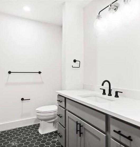 A modern bathroom with white walls and a black hexagonal tile floor. It features a large white vanity with a sink, black fixtures, a toilet, and a black towel rack. The lighting fixture has three round bulbs above the vanity.