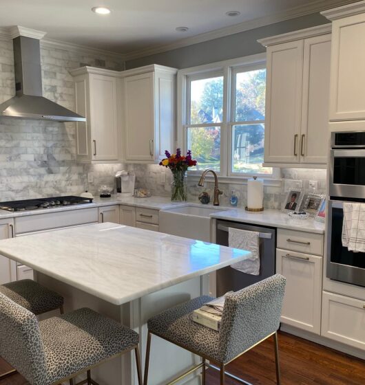 A modern kitchen with white cabinets, marble countertops, and a central island. It features a gas stove with a stainless steel hood, built-in double ovens, and a vase of flowers near the sink. Four patterned stools surround the island.